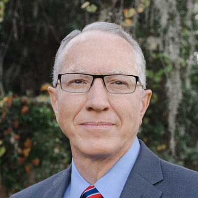 Dr. Keatley Waldron wearing glasses, a suit, and a striped tie, photographed outdoors with trees